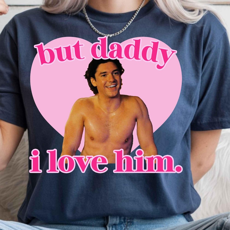 Puede incluir: Una camiseta azul oscuro con un gr&aacute;fico de un coraz&oacute;n rosa grande que contiene la fotograf&iacute;a de un hombre sin camisa. El texto "but daddy i love him." est&aacute; impreso en letras rosas sobre y debajo del coraz&oacute;n en una fuente negrita.