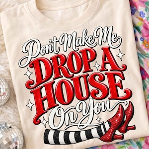 May include: A cream-colored t-shirt featuring the text "Don't Make Me DROP A HOUSE On You" in black and red glittery lettering. The design includes small star accents and a graphic of two legs wearing black and white striped stockings and red heels.