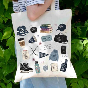 Puede incluir: Una bolsa de mano blanca con ilustraciones coloridas sobre el tema universitario, que incluyen una camiseta de hockey, libros, una taza de caf&eacute; y una botella de agua. La bolsa tiene el texto "Briar U" y "#1 FAN".