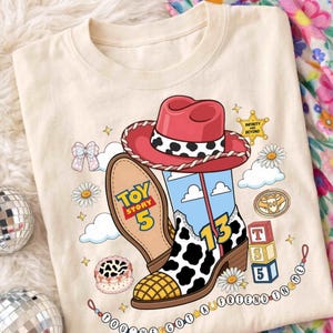 May include: A cream-colored t-shirt featuring a graphic of a red cowboy hat and a cow-print boot. The design includes the text Toy Story 5, the number 13, and You've Got A Friend In Me, alongside illustrations of daisies, clouds, and blocks.