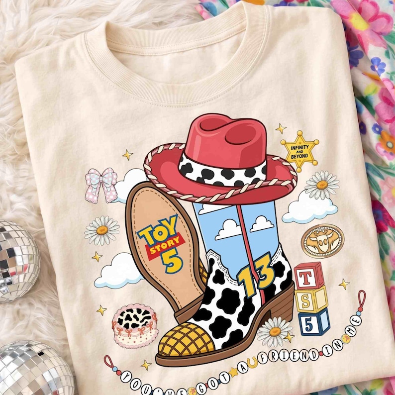 May include: A cream-colored t-shirt featuring a graphic of a red cowboy hat and a cow-print boot. The design includes the text Toy Story 5, the number 13, and You've Got A Friend In Me, alongside illustrations of daisies, clouds, and blocks.
