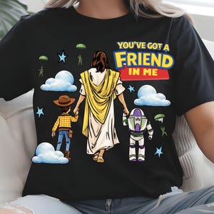 May include: Black t-shirt featuring a graphic of a figure in a yellow robe holding hands with two cartoon characters. The text "YOU'VE GOT A FRIEND IN ME" is displayed in colorful letters above the figures. Stars, clouds, and parachuters are also in the design.