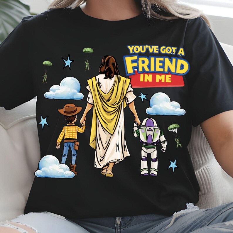 May include: Black t-shirt featuring a graphic of a figure in a yellow robe holding hands with two cartoon characters. The text "YOU'VE GOT A FRIEND IN ME" is displayed in colorful letters above the figures. Stars, clouds, and parachuters are also in the design.