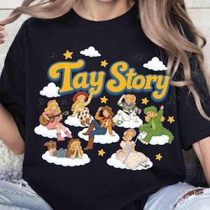 May include: A black t-shirt featuring the yellow text Tay Story. The graphic includes seven illustrated characters with blonde hair, each sitting on a white cloud, surrounded by yellow stars, white clouds, and black musical note symbols.
