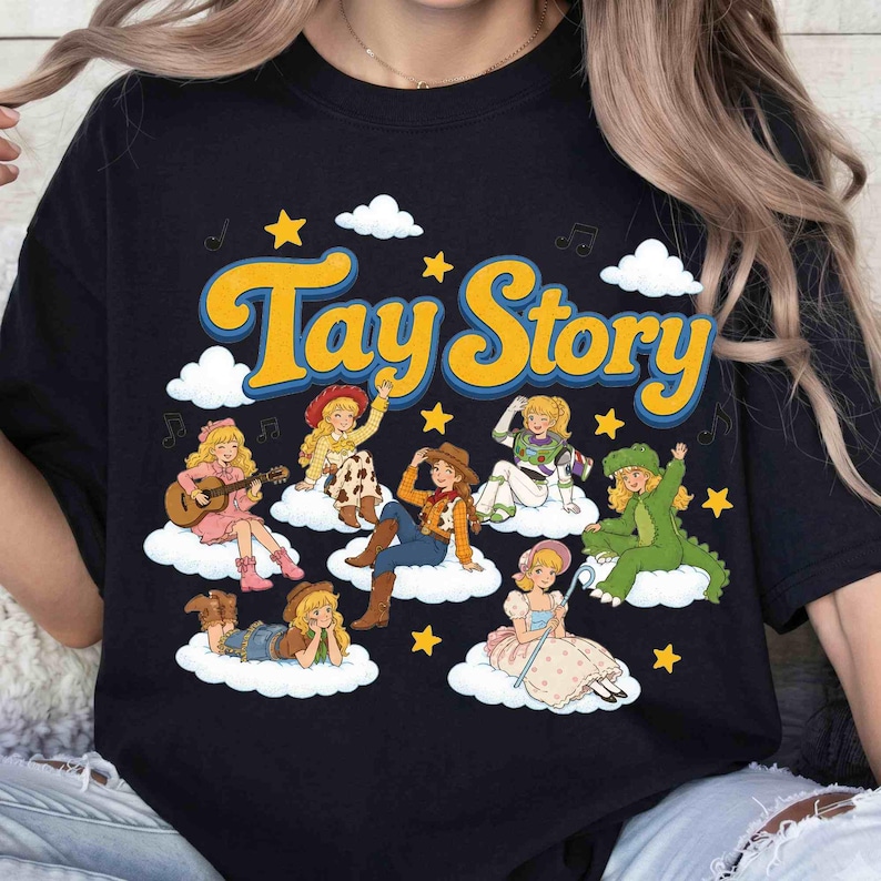 May include: A black t-shirt featuring the yellow text Tay Story. The graphic includes seven illustrated characters with blonde hair, each sitting on a white cloud, surrounded by yellow stars, white clouds, and black musical note symbols.