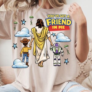 May include: Beige t-shirt with a graphic featuring Jesus holding hands with Woody and Buzz Lightyear. The text "YOU'VE GOT A FRIEND IN ME" is displayed above the figures, along with clouds, stars, and parachuters.