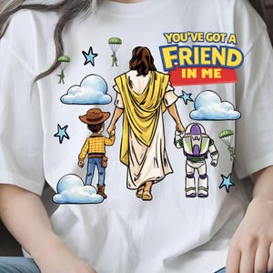 May include: White t-shirt featuring a graphic design with the text "YOU'VE GOT A FRIEND IN ME". The design includes cartoon illustrations of Jesus, Woody, and Buzz Lightyear, along with clouds, stars, and parachuting figures.