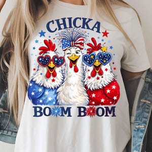 Puede incluir: Camiseta blanca con un dise&ntilde;o de tres pollos de dibujos animados con colores patri&oacute;ticos. Los pollos llevan gafas de sol en forma de coraz&oacute;n y tienen detalles en rojo, blanco y azul. El texto "CHICKA BOOM BOOM" est&aacute; impreso en azul.