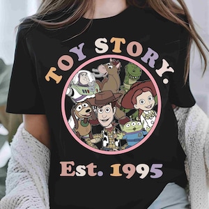 May include: A black t-shirt featuring a circular graphic with Toy Story characters including Woody, Buzz Lightyear, Jessie, Rex, Bullseye, Slinky Dog, and an Alien. The text reads TOY STORY and Est. 1995 in colorful, retro-style lettering.