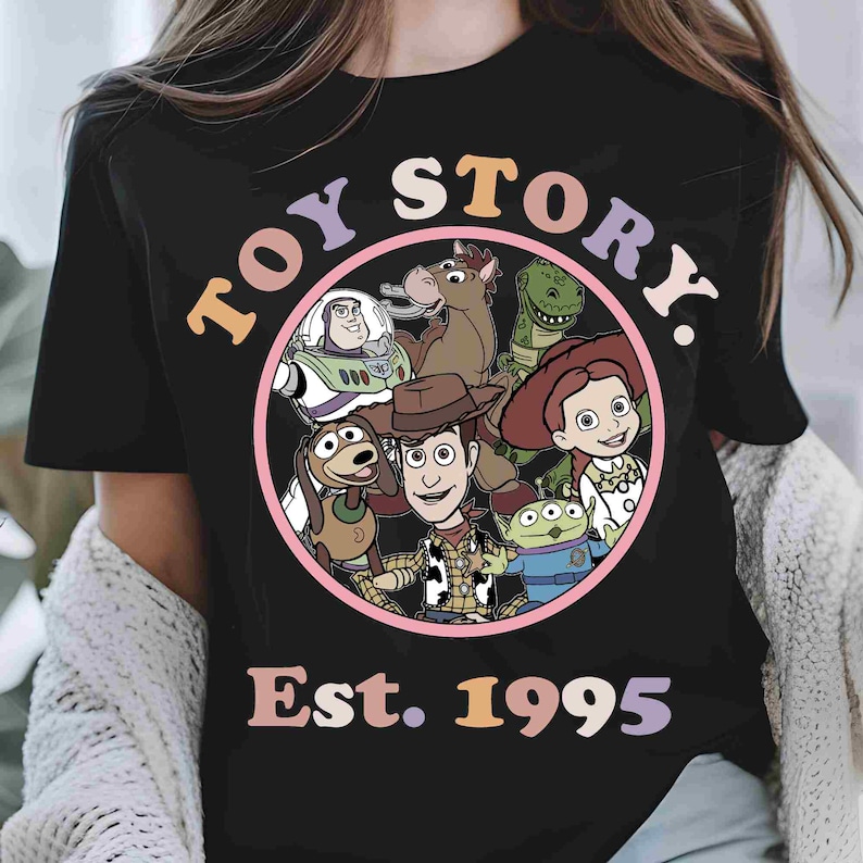 May include: A black t-shirt featuring a circular graphic with Toy Story characters including Woody, Buzz Lightyear, Jessie, Rex, Bullseye, Slinky Dog, and an Alien. The text reads TOY STORY and Est. 1995 in colorful, retro-style lettering.