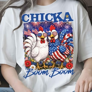 Puede incluir: Camiseta blanca con un gr&aacute;fico de dos gallos con gafas de sol con el dise&ntilde;o de la bandera estadounidense. La palabra "CHICKA" est&aacute; encima de los gallos, y "Boom Boom" est&aacute; escrito debajo. Fuegos artificiales y banderas americanas en el fondo.
