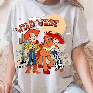 May include: A light gray t-shirt featuring a graphic print of a cowboy, a cowgirl, and a brown horse in a desert landscape. The text WILD WEST is printed in orange above the characters, with cacti, clouds, and a large sun in the background.