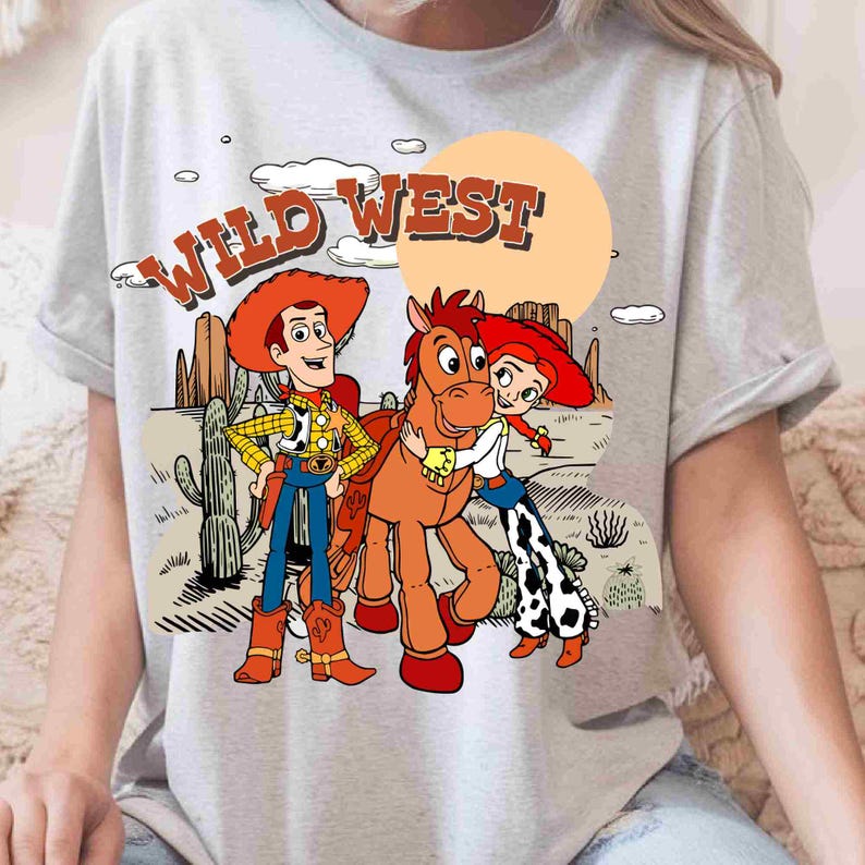 May include: A light gray t-shirt featuring a graphic print of a cowboy, a cowgirl, and a brown horse in a desert landscape. The text WILD WEST is printed in orange above the characters, with cacti, clouds, and a large sun in the background.