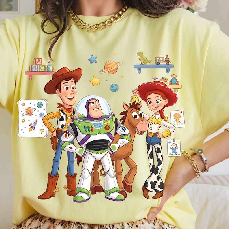 May include: A pale yellow t-shirt featuring a colorful graphic print of Woody, Buzz Lightyear, Bullseye, and Jessie from Toy Story. The design includes floating planets, stars, and small shelves with toys, set against a light yellow fabric background.