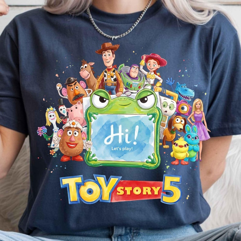 May include: A navy blue t-shirt featuring a graphic print of various Toy Story characters surrounding a green frog-shaped frame with the text Hi! Let's play! inside. Below the characters, the text TOY STORY 5 is printed in large yellow letters.