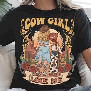 May include: A black t-shirt featuring a graphic illustration of two figures in cowboy hats standing in a desert landscape with cacti and roses. The text COW GIRL is arched above, and LIKE ME is on a banner below, with I KNEW IT written in circles.