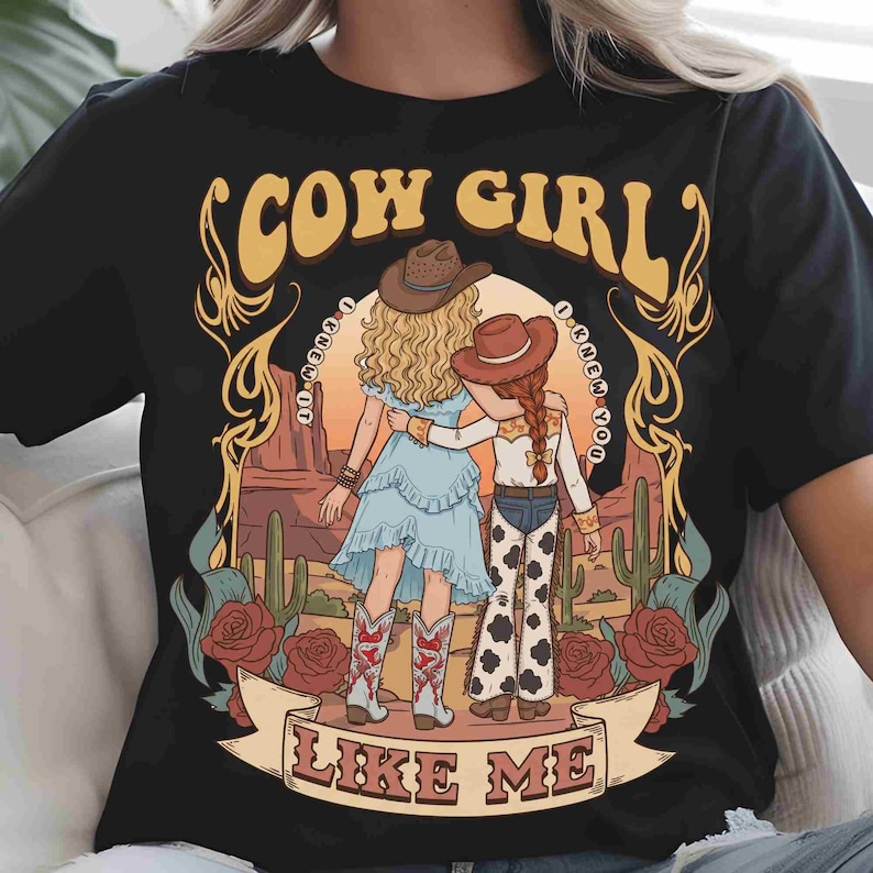 May include: A black t-shirt featuring a graphic illustration of two figures in cowboy hats standing in a desert landscape with cacti and roses. The text COW GIRL is arched above, and LIKE ME is on a banner below, with I KNEW IT written in circles.