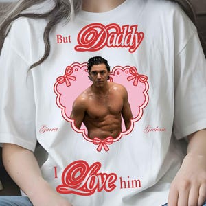 Puede incluir: Una camiseta blanca con un estampado gr&aacute;fico rojo que dice But Daddy I Love him. El dise&ntilde;o incluye un marco rosa en forma de coraz&oacute;n con lazos rojos que contiene la foto de un hombre sin camisa, con los nombres Garret y Graham en rojo.