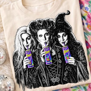 May include: A cream-colored t-shirt featuring a black and white graphic of three figures with elaborate hairstyles, each holding a purple Alani energy drink can. The graphic includes detailed line work and text on the cans reading Alani.