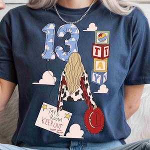 May include: A navy blue t-shirt featuring a graphic of a person with long blonde hair, a cow-print jacket with red fringe, and a red hat. The design includes a number 13 balloon, stacked alphabet blocks, and a sign reading Tays Room Keep Out.