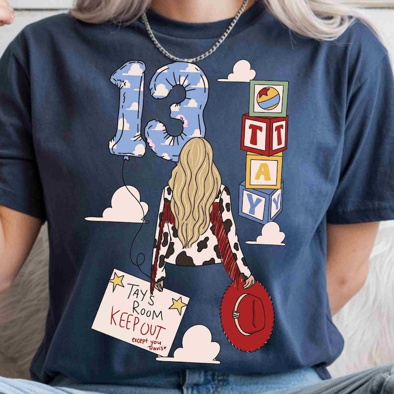 May include: A navy blue t-shirt featuring a graphic of a person with long blonde hair, a cow-print jacket with red fringe, and a red hat. The design includes a number 13 balloon, stacked alphabet blocks, and a sign reading Tays Room Keep Out.