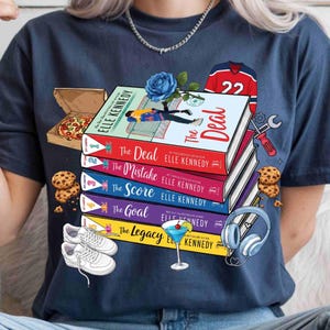 May include: A navy blue t-shirt with a design featuring a stack of books titled "The Deal", "The Mistake", "The Score", "The Goal", and "The Legacy" by Elle Kennedy. The design includes pizza, cookies, trainers, and a cocktail.