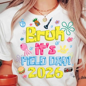 Op de afbeelding: Wit T-shirt met een kleurrijk grafisch ontwerp. Het ontwerp bevat de woorden "Bruh It's Field Day! 2026" in geel, roze en blauw, samen met cartoon illustraties van een ananas, klarinet en andere speelse elementen.