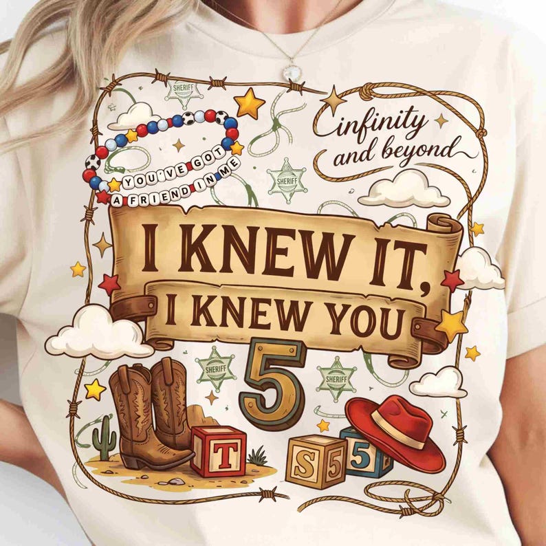 May include: A cream-colored t-shirt featuring a graphic print with the text "Cinfinity and beyond", "I KNEW IT, I KNEW YOU", and "YOU'VE GOT A FRIEND IN ME". The design includes cowboy boots, a red hat, alphabet blocks, sheriff stars, and a lasso.