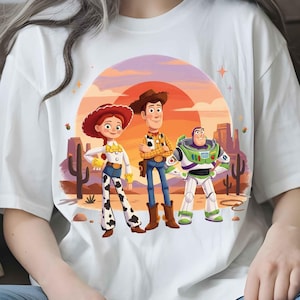 May include: A white t-shirt featuring a graphic print of three animated characters: a cowgirl with a red hat, a cowboy with a brown hat, and a space ranger in a white and green suit. The background shows a desert landscape with cacti and a sunset.