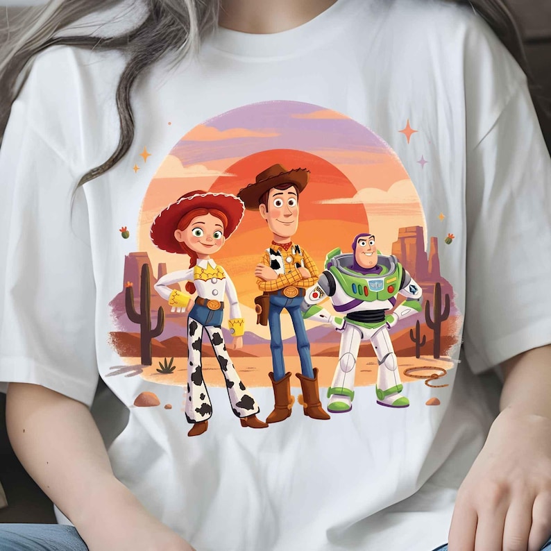 May include: A white t-shirt featuring a graphic print of three animated characters: a cowgirl with a red hat, a cowboy with a brown hat, and a space ranger in a white and green suit. The background shows a desert landscape with cacti and a sunset.