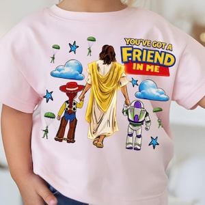 May include: A light pink t-shirt featuring a graphic with the text "YOU'VE GOT A FRIEND IN ME". The design includes illustrations of Jesus, Woody, and Buzz Lightyear, along with clouds, stars, and parachuters.