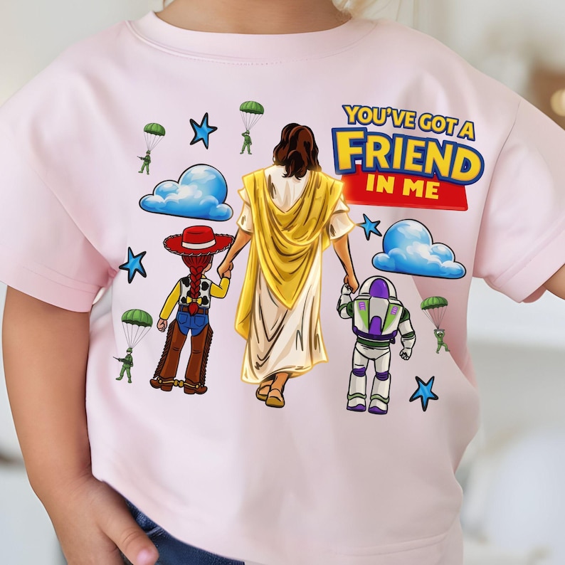 May include: A light pink t-shirt featuring a graphic with the text "YOU'VE GOT A FRIEND IN ME". The design includes illustrations of Jesus, Woody, and Buzz Lightyear, along with clouds, stars, and parachuters.