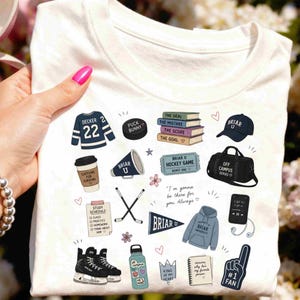 Puede incluir: Camiseta blanca con varias ilustraciones gr&aacute;ficas, incluyendo una camiseta de hockey, un disco, libros, un gorro, una taza de caf&eacute;, palos de hockey, una botella de agua y un dedo de espuma. La camiseta tiene el texto "Briar U" y otras frases.