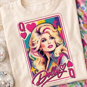 May include: A cream-coloured t-shirt featuring a graphic of a playing card with a blonde woman, pink hearts, and yellow stars. The text Dolly is written in a cursive script at the bottom, with the letter Q in the corners of the rectangular frame.