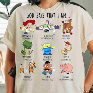 May include: A cream-colored t-shirt featuring the text GOD SAYS THAT I AM... above nine cartoon characters from Toy Story, each paired with a descriptive word and a Bible verse reference, including Jessie, Buzz Lightyear, Bullseye, Rex, Alien, Woody, Slinky Dog, Mr. Potato Head, and Hamm.
