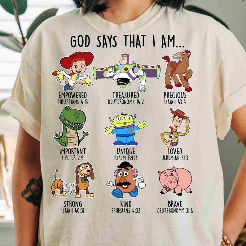 May include: A cream-colored t-shirt featuring the text GOD SAYS THAT I AM... above nine cartoon characters from Toy Story, each paired with a descriptive word and a Bible verse reference, including Jessie, Buzz Lightyear, Bullseye, Rex, Alien, Woody, Slinky Dog, Mr. Potato Head, and Hamm.