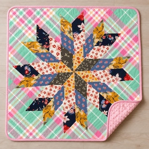 Könnte beinhalten: Ein quadratischer Quilt mit einem zentralen Sternmuster aus verschiedenen floralen und geometrischen Stoffstücken in Marineblau, Senfgelb, Weiß und Rot. Der Hintergrund hat ein diagonales Karomuster in Mintgrün und Pink mit einem rosa Rand.