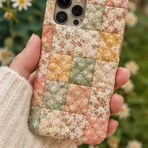 May include: A quilted phone case featuring a patchwork design of floral patterns in shades of pink, yellow, and green. The case has a square-stitched texture and includes the text "Soft, Quilted & Durable" printed on a pink rectangular banner at the bottom.