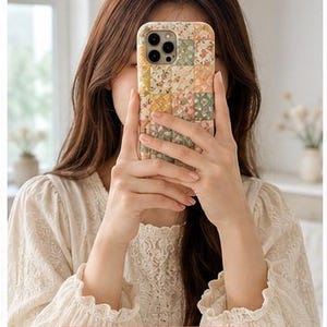 May include: A rectangular smartphone case featuring a patchwork design of various floral patterns in soft shades of yellow, green, pink, and white. The case has a camera cutout at the top left and a rectangular pink label at the bottom with the text Handmade with Love and a white heart symbol.