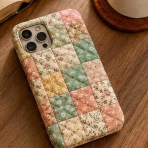 May include: A rectangular phone case featuring a quilted patchwork design with a grid of squares in soft shades of pink, green, yellow, and cream, each decorated with a small floral pattern. The case has a cutout for a triple camera lens system.