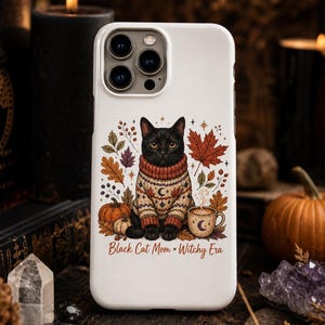 Puede incluir: Funda de teléfono blanca con un gato negro con un suéter, rodeado de hojas de otoño, calabazas y una taza de café. El texto "Black Cat Mom Witchy Era" está impreso debajo.