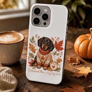 Puede incluir: Funda de teléfono blanca con un diseño de perro salchicha con un suéter, rodeado de hojas de otoño. La funda incluye el texto "Dachshund Mom • Long Dog Lover" y una taza de café con un corazón. También se ven una calabaza y una taza de café.