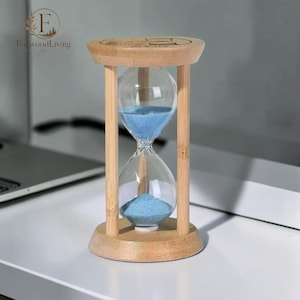 May include: A wooden hourglass with light blue sand. The hourglass is made of clear glass and is held in place by a light brown wooden frame. The hourglass is on a white surface.