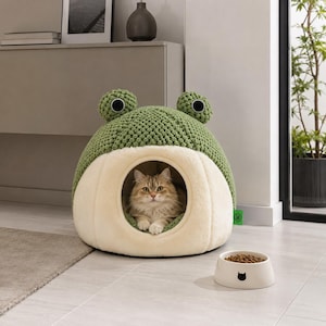Puede incluir: Una cama para mascotas con forma de rana en color verde y crema, con una parte superior de punto texturizado y una abertura frontal de felpa. Un gato de pelo largo descansa dentro. Un cuenco redondo color crema con un icono de gato negro.