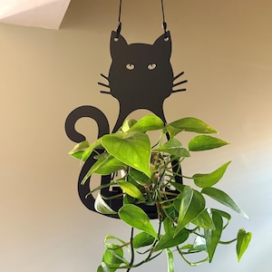 May include: A black metal hanging planter shaped like a cat silhouette with a curled tail. The planter features a flat, cutout design with two small holes at the top for suspension cords, holding a trailing green plant with heart-shaped leaves.