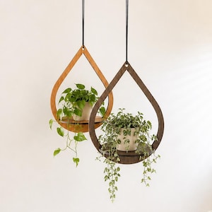 May include: Two teardrop-shaped wooden hanging planters suspended by black cords. One frame is light brown wood, the other is dark brown wood. Each holds a white pot containing a green trailing plant with heart-shaped leaves.