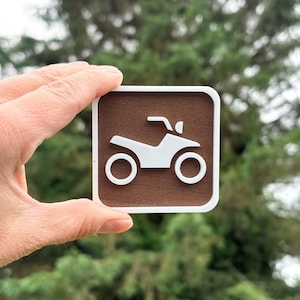 Peut inclure: Un panneau carré avec un fond texturé marron foncé, une bordure blanche et un graphique en relief blanc représentant un véhicule tout-terrain ou quad au centre. Le panneau présente des coins arrondis et un design contrasté.