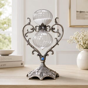 May include: An ornate hourglass with a clear glass bulb and white sand. The hourglass is supported by a decorative metal frame with a floral base and a blue gem. The design is vintage and elegant.