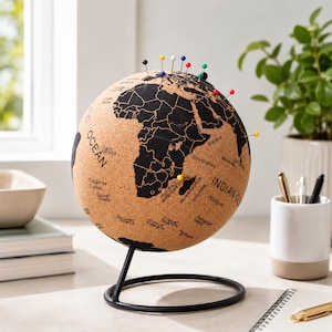 May include: A cork globe with black continents and country names, mounted on a black metal stand. Various coloured pins mark locations. The globe is on a white surface with a plant, pens, and a notebook in the background.
