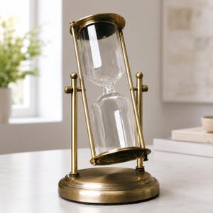 May include: A brass-coloured hourglass with a clear glass central section. The hourglass is tilted, showing sand flowing downwards. The base and top are circular, and the frame has a classic design. The height is approximately 20 cm.